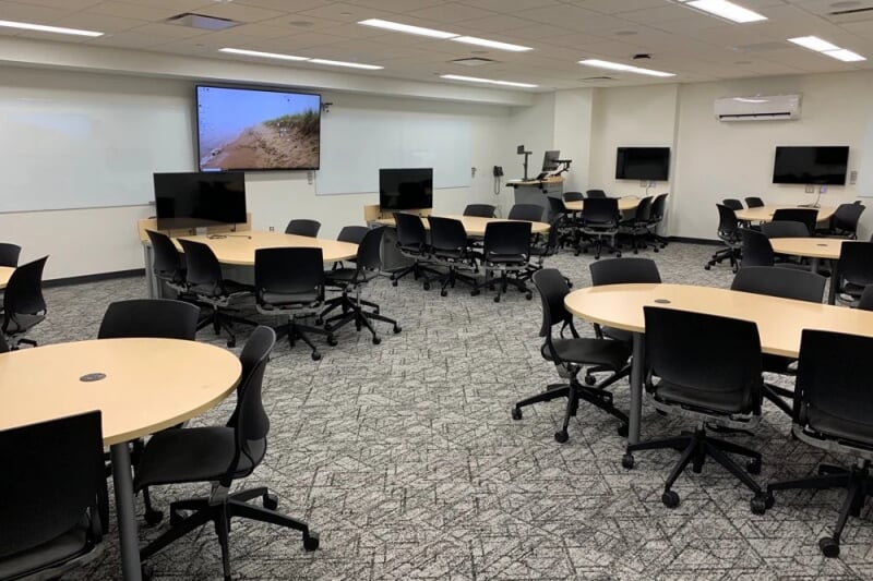 Chem-Sci B002 showing 8 group tables surrounded by chairs with casters, each with a monitor mounted on the wall nearby. A large monitor and two large white boards are on the wall.