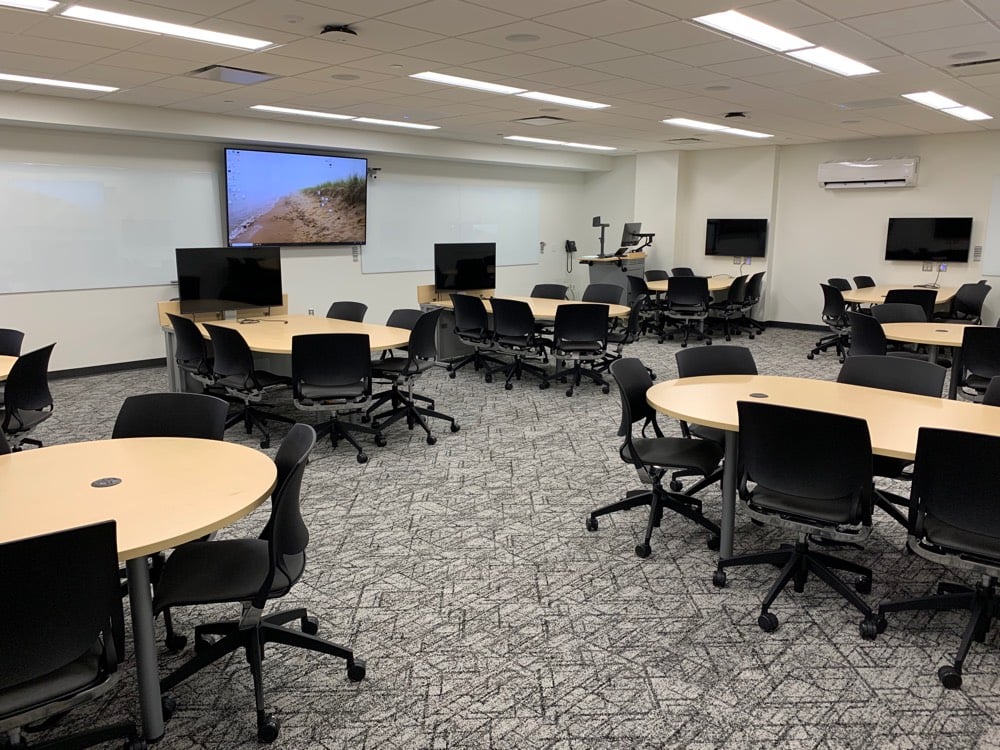 Chem-Sci B002 showing 8 group tables surrounded by chairs with casters, each with a monitor mounted on the wall nearby. A large monitor and two large white boards are on the wall.