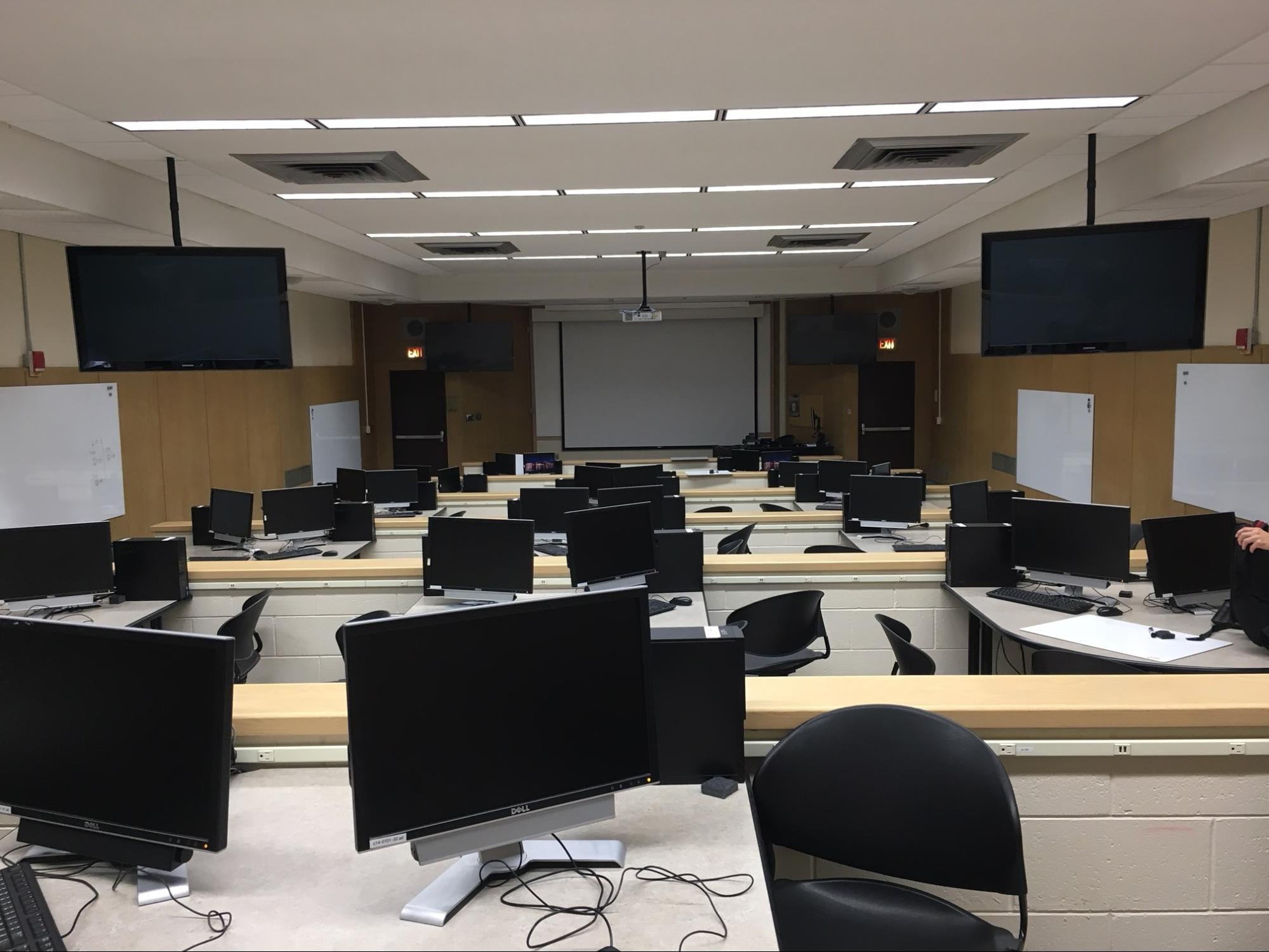 Dillman 101 is a long tiered classroom with multiple small group tables per tier, each table having two computers. Several monitors hang from the ceiling and white boards are mounted along the walls.