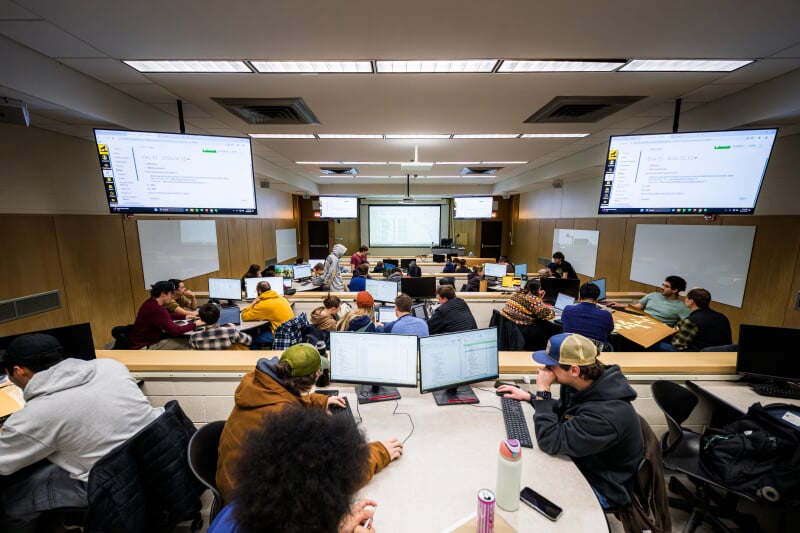 Dillman 101 is a long tiered classroom with multiple small group tables per tier, each table having two computers. Several monitors hang from the ceiling and white boards are mounted along the walls.