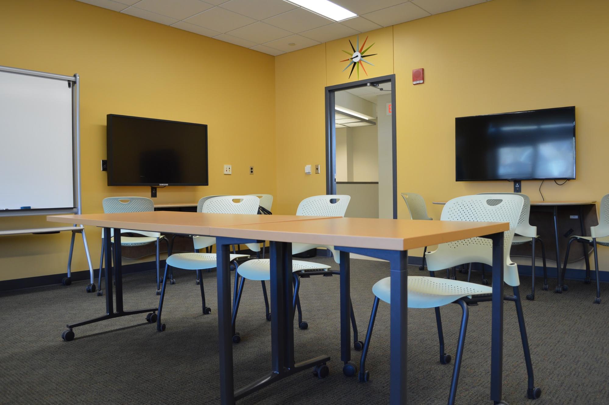 Library 243 showing a narror table with 3 chairs, all on wheels. Two large monitors and a mobile white board are against the walls.