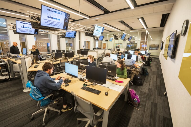MEEM 120 showing tables with 4 computer stations each and monitors mounted both on the walls and from trusses near the ceiling.