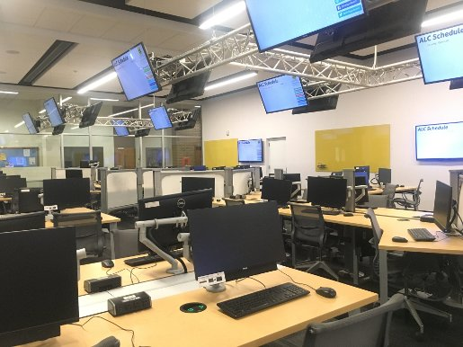 MEEM 120 showing tables with 4 computer stations each and monitors mounted both on the walls and from trusses near the ceiling.