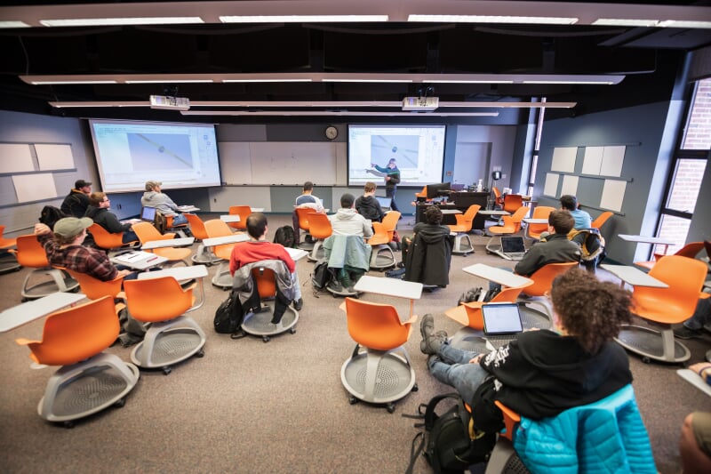 MEEM 402 showing a number of free range tablet-arm chairs with casters spread throughout the room. Dual projectors with one agaist the wall and one in the corner and multiple removable white boards on the walls.