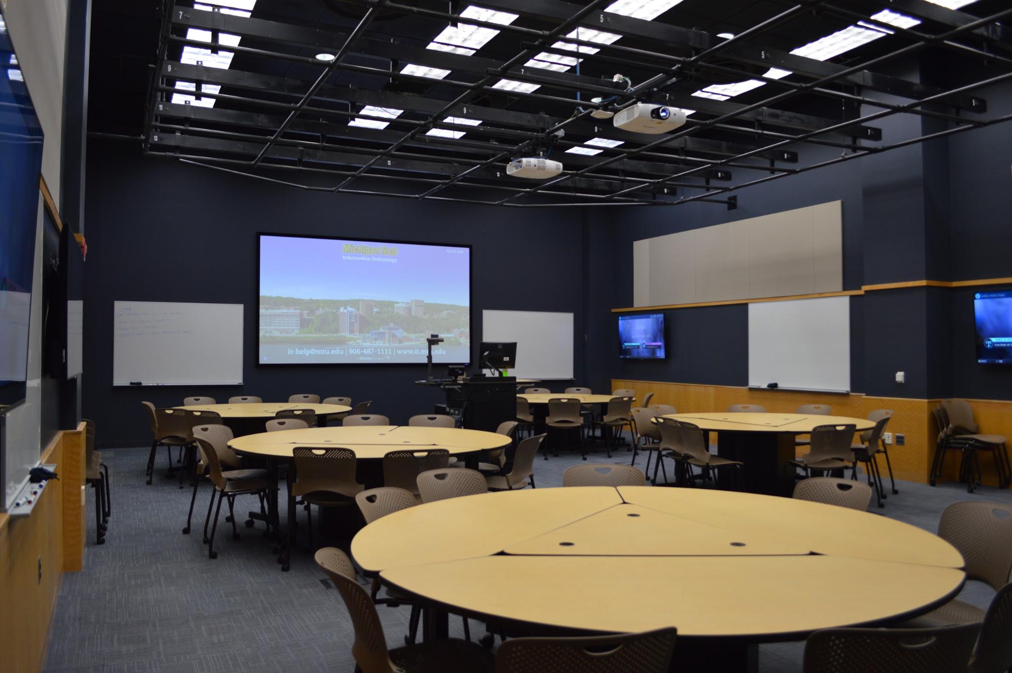 Rekhi G05 - five round tables in a room with a projector and several monitors and white boards around the edge of the room.