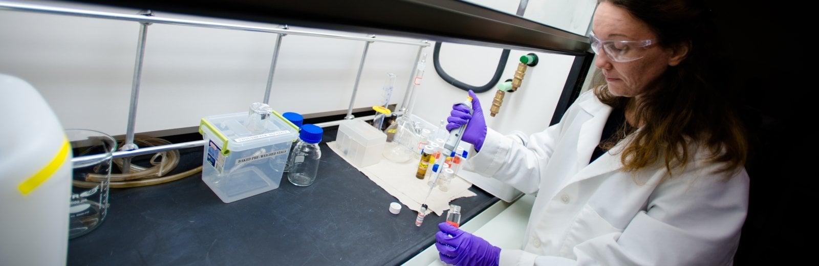 Researcher in lab with gloves and safety goggle under a chemical hood.