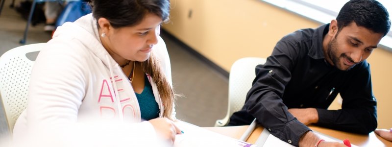 Two learning center students sitting, one is writing with a dry erase marker while the other looks on with an open book.