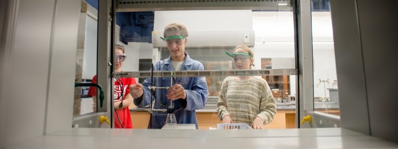 Three students using an Erlenmeyer flask inside a fume hood