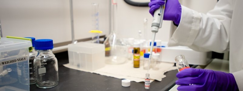 A chemist in purple gloves transfers a liquid from one vial to another using a pipet