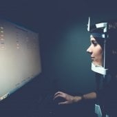 Participant sitting with head in a device that tracks eye movement while looking at a monitor.