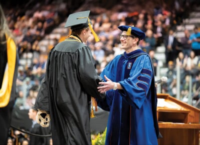 Dennis Livesay shaking hands with a student wearing Blizzard mascot paws during commencement.