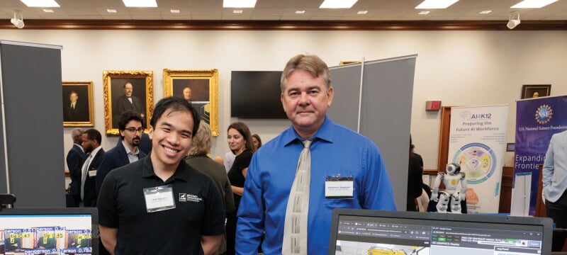 Vinh Nguyen and Alex Sergeyev stand behind a table with two computer monitors.
