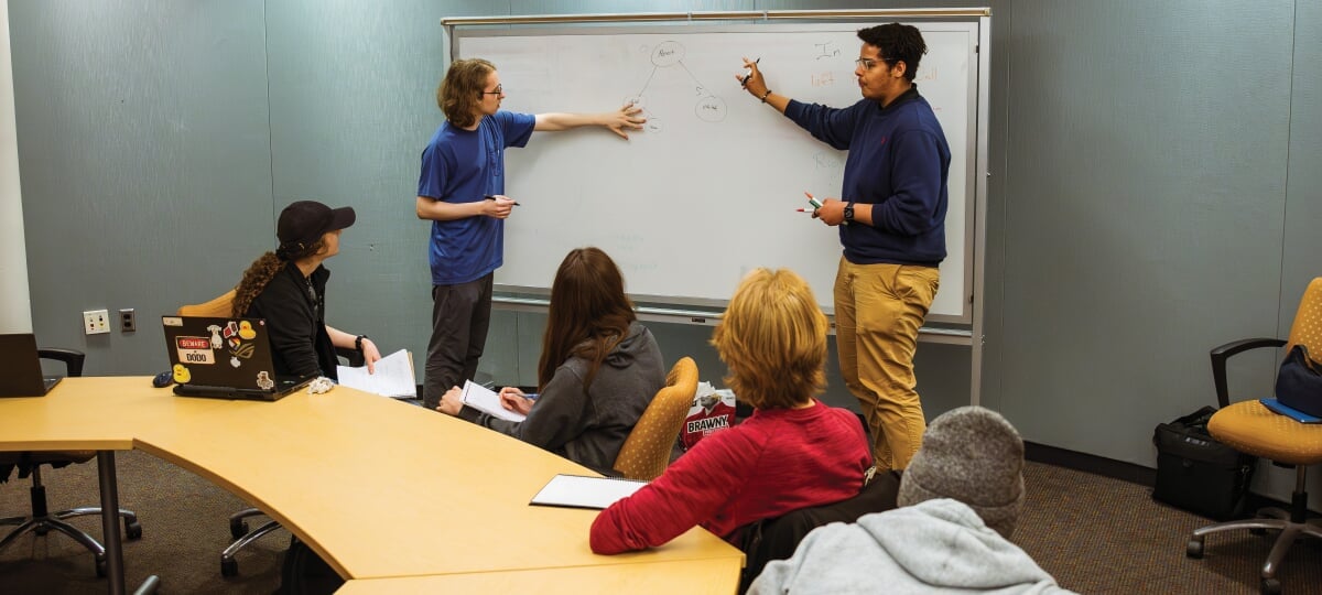 Two students working at a whiteboard while others look on.