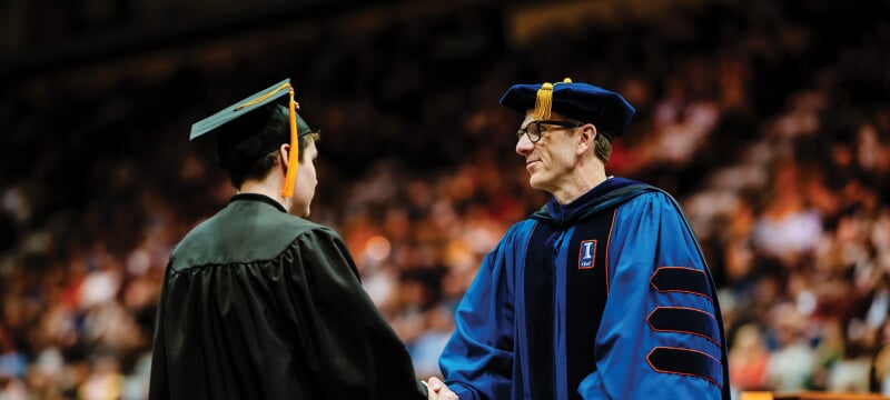 Dean Livesay, in full regalia, shakes hands with a student graduate at commencement.