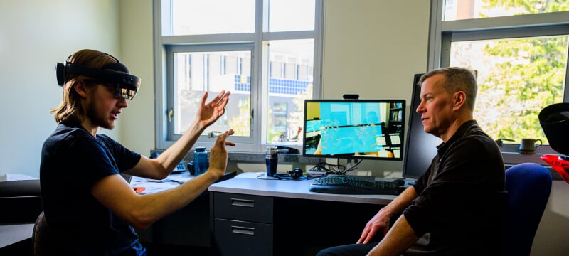 Student wearing a virtual reality headset sitting next to a faculty member at a desk.