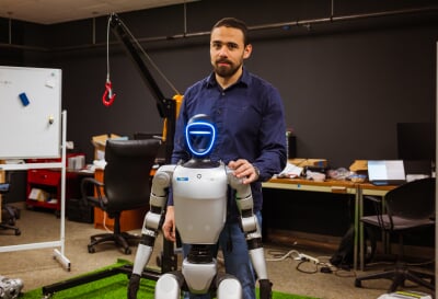 Person standing behind a humanoid robot with his hand on the shoulder.