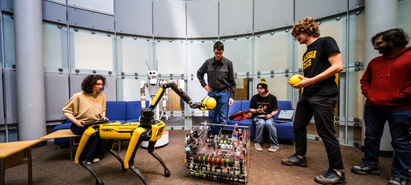Students in a lounge with several robots including a humanoid-looking one, a dog looking one with an arm holding a ball, and a rectangular one.