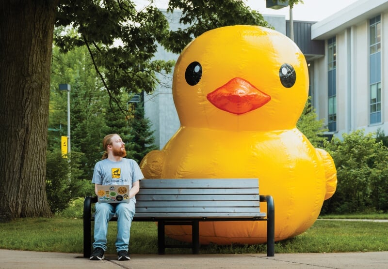 Gabrial Belanger sitting on a bench with a laptop looking over his shoulder at a giant blow-up duck.