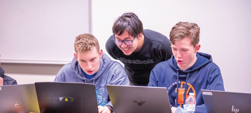 One student looking over the shoulders of two others working on laptops.