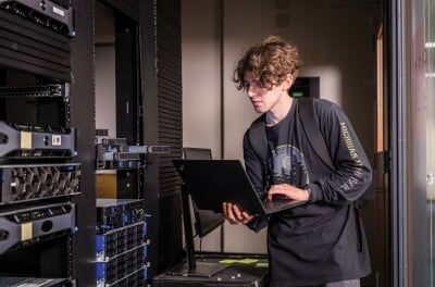 Student holding a laptop looking at a server rack.