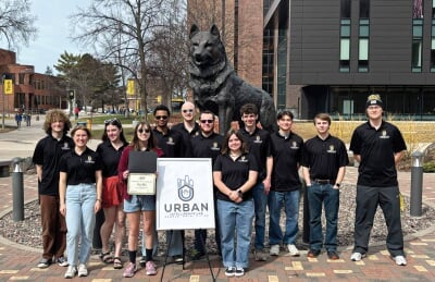 UI Lab Enterprise group standing in front of the Husky Statue with a sign featuring their logo.