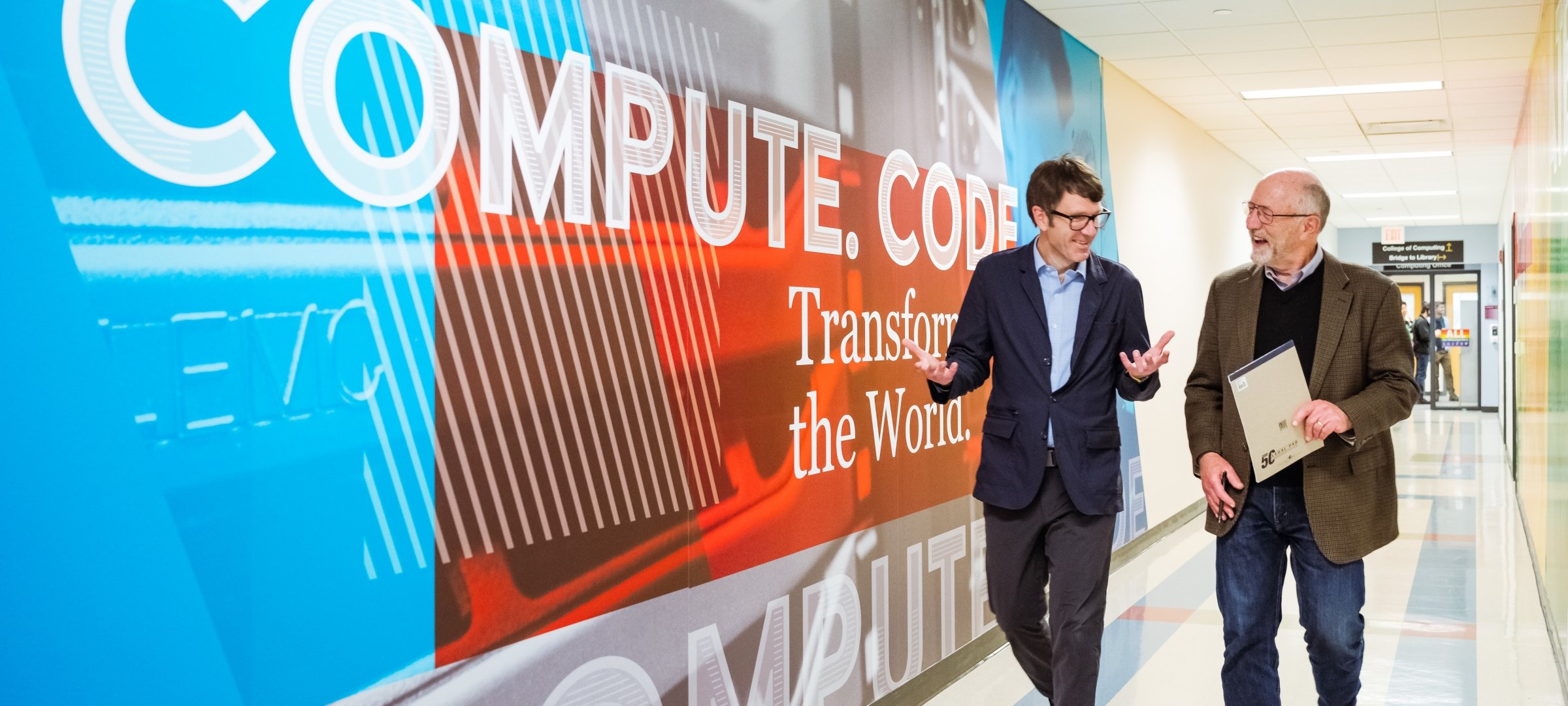 Two people walking down a hall with the words Compute Code Transform the World written on the wall