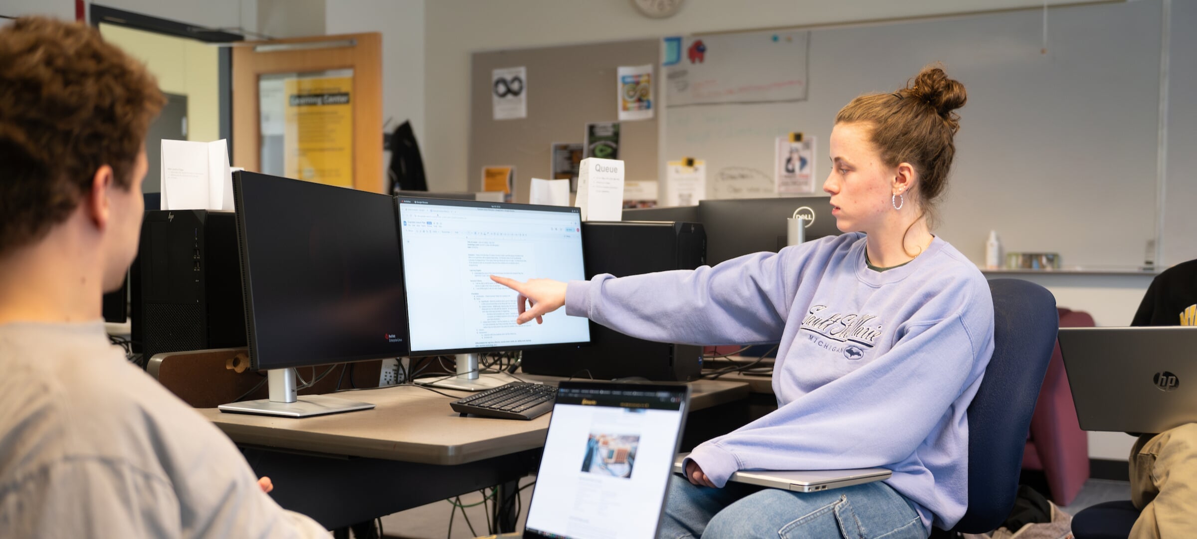 Two students in a lab, one pointing to a screen, the other looking on with a laptop.