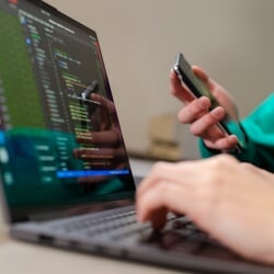 Hands typing on a laptop displaying code while another person holds a smartphone.