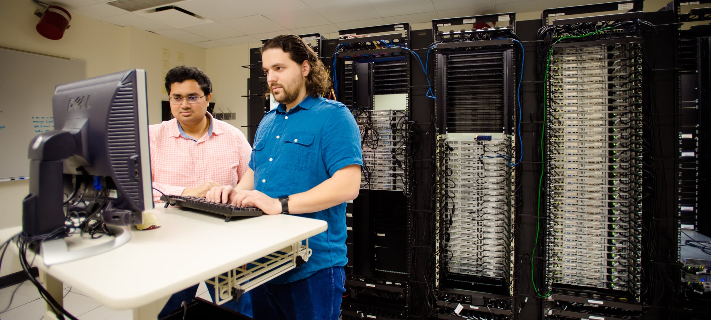 Two graduate students standing in front of a computer with a wall of servers behind them.
