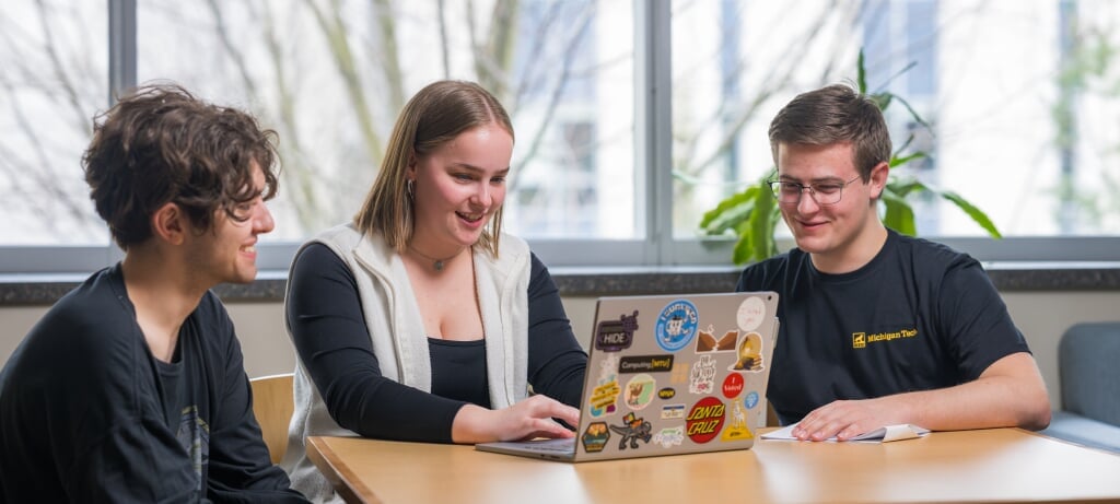 Three Michigan Tech College of Computing students hanging out in the college lounge looking at a laptop with plants, windows, and a campus view behind them.