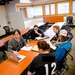 Students sitting at a table with a professor looking at a laptop.