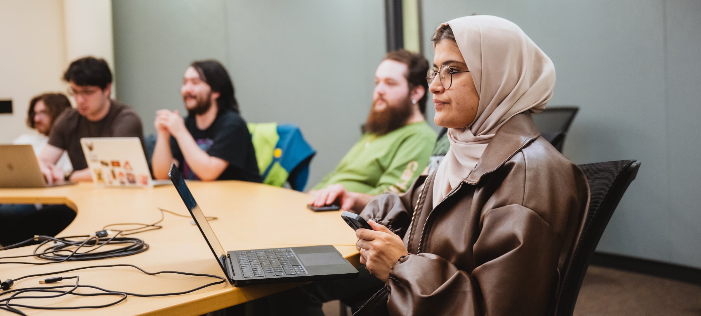 Students sit at a curved table with laptops and phones out, listening to an off-screen speaker.