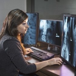 A student sits at a table, examining different x-rays on three computer monitors.