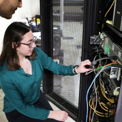 A student crouches beside an open server, inspecting the wires within.