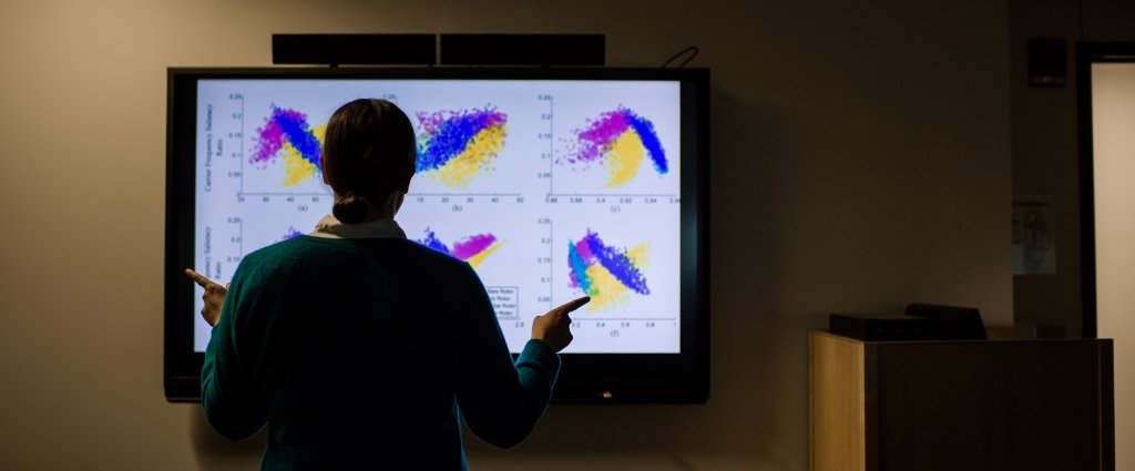 Faculty standing in front of a monitor pointing to a grid of charts