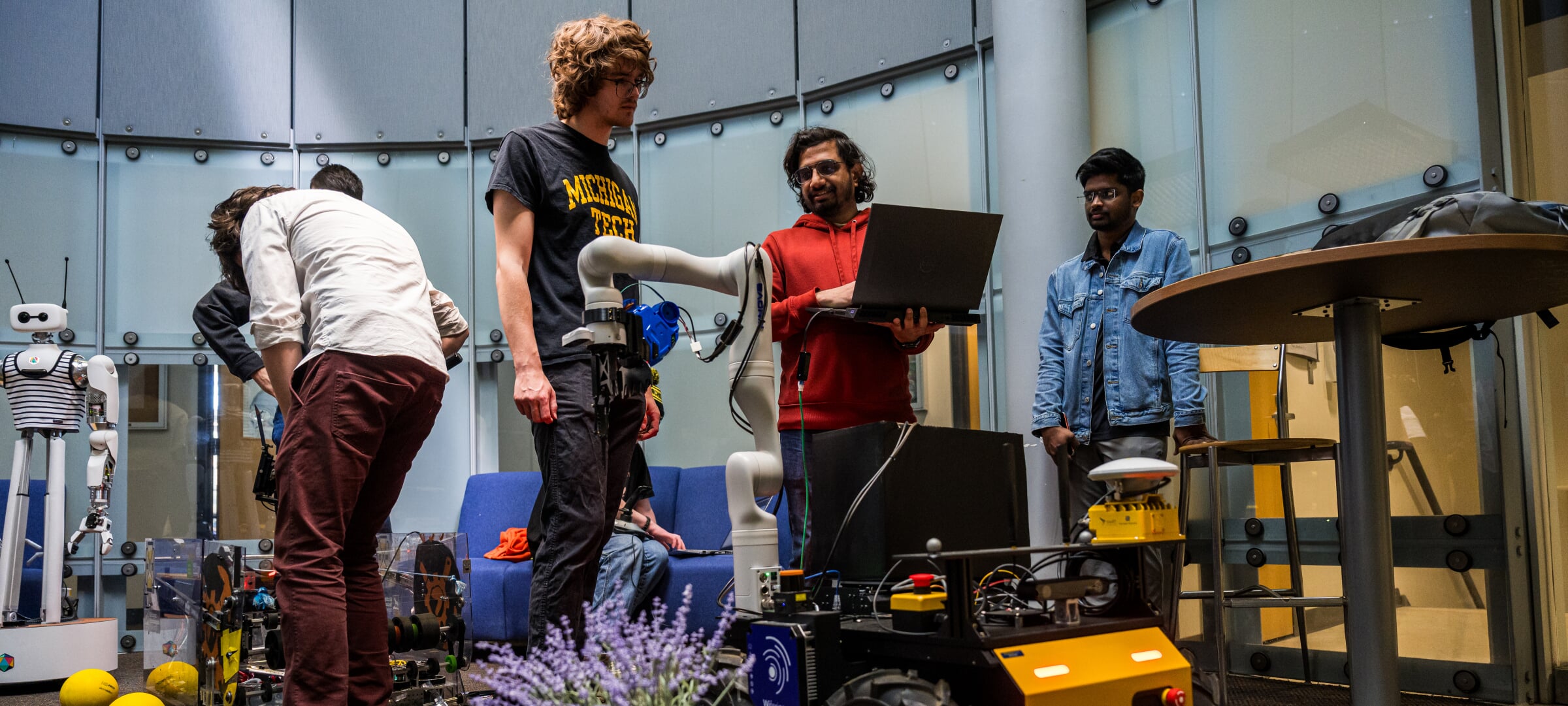 A group of students, with one holding a laptop, around a robot talking.