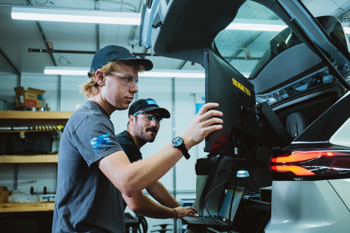 two people working on a computer next to a car inside