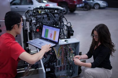 Two students inspect an internal combustion engine using a laptop hooked into the system.