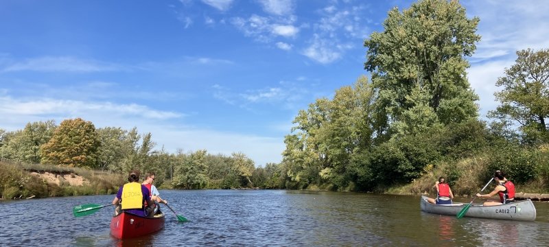 4 students split between two canoes paddling on a river surrounded by trees