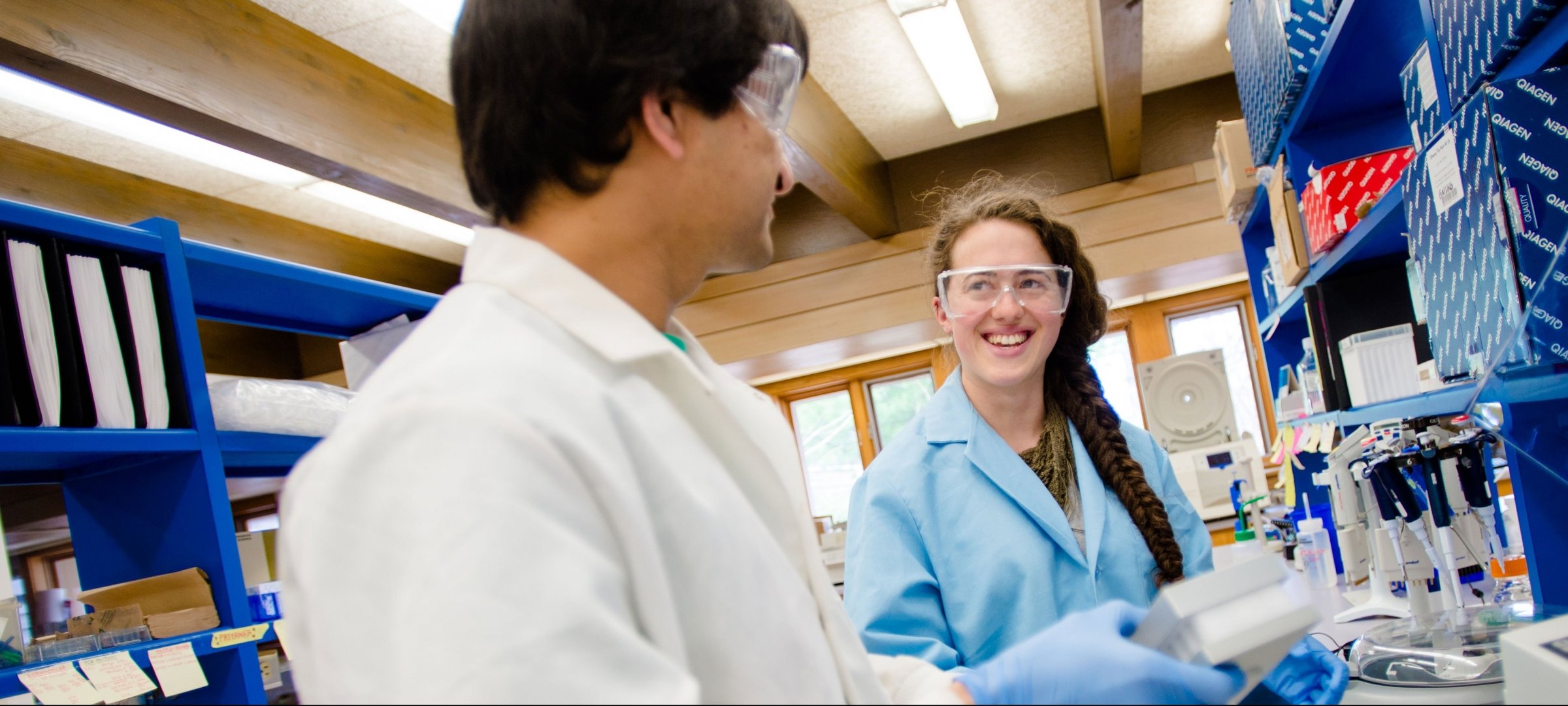Two graduate students inside a lab with lab coats, gloves, and safety glasses.