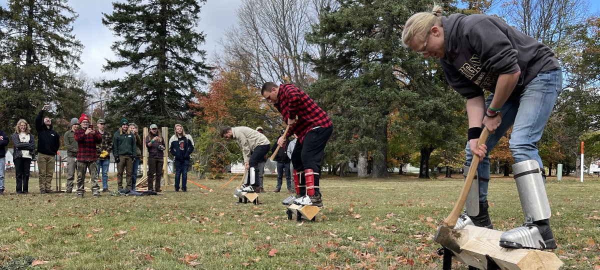 Three people stand on logs on stands chopping while spectators look on as Society of American Foresters Forestry Club students from Michigan Tech and other colleges compete.