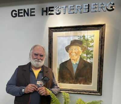 RJ Laverne holding a compass and standing next to a portrait of Gene Hesterberg.