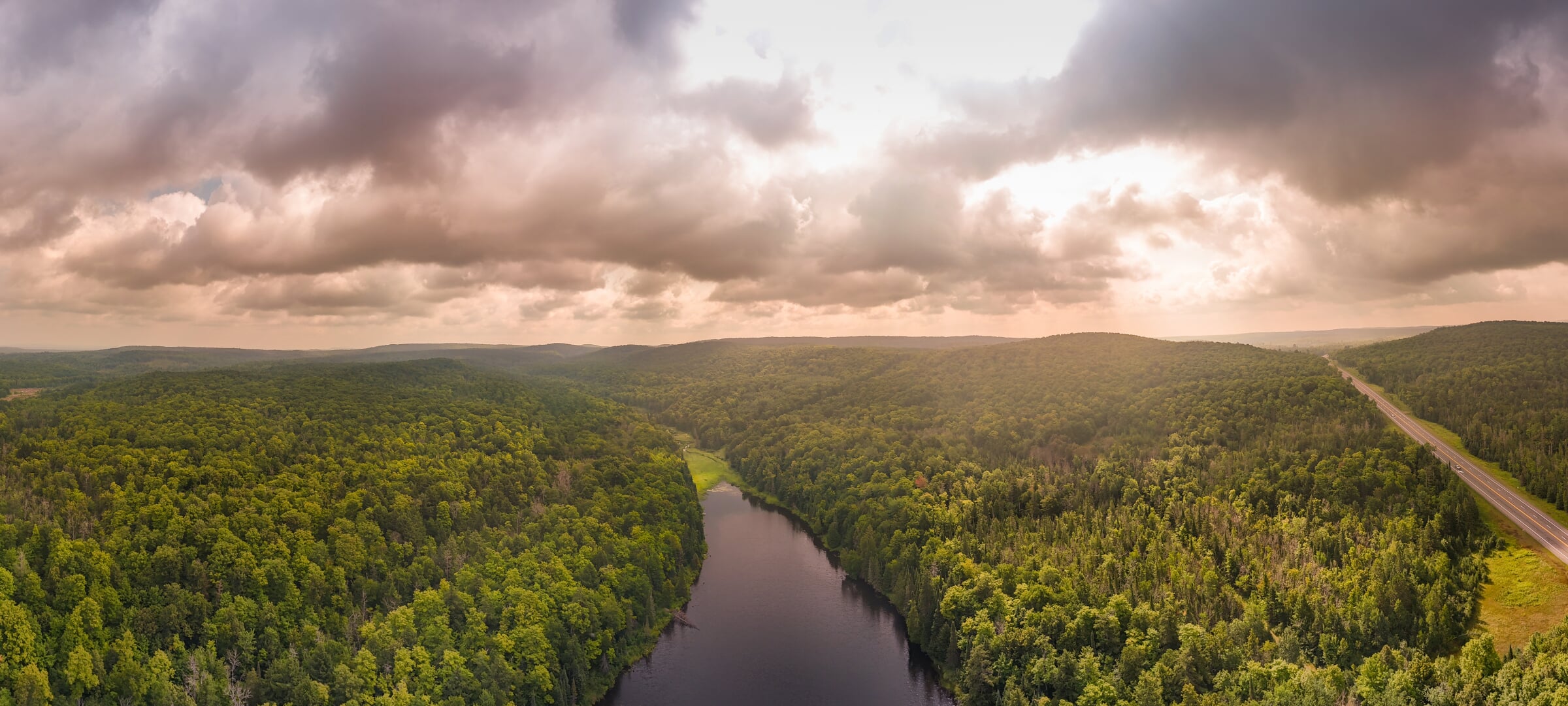 Aerial view of a large forest, a river through it, and a road to the right, with the sun setting.