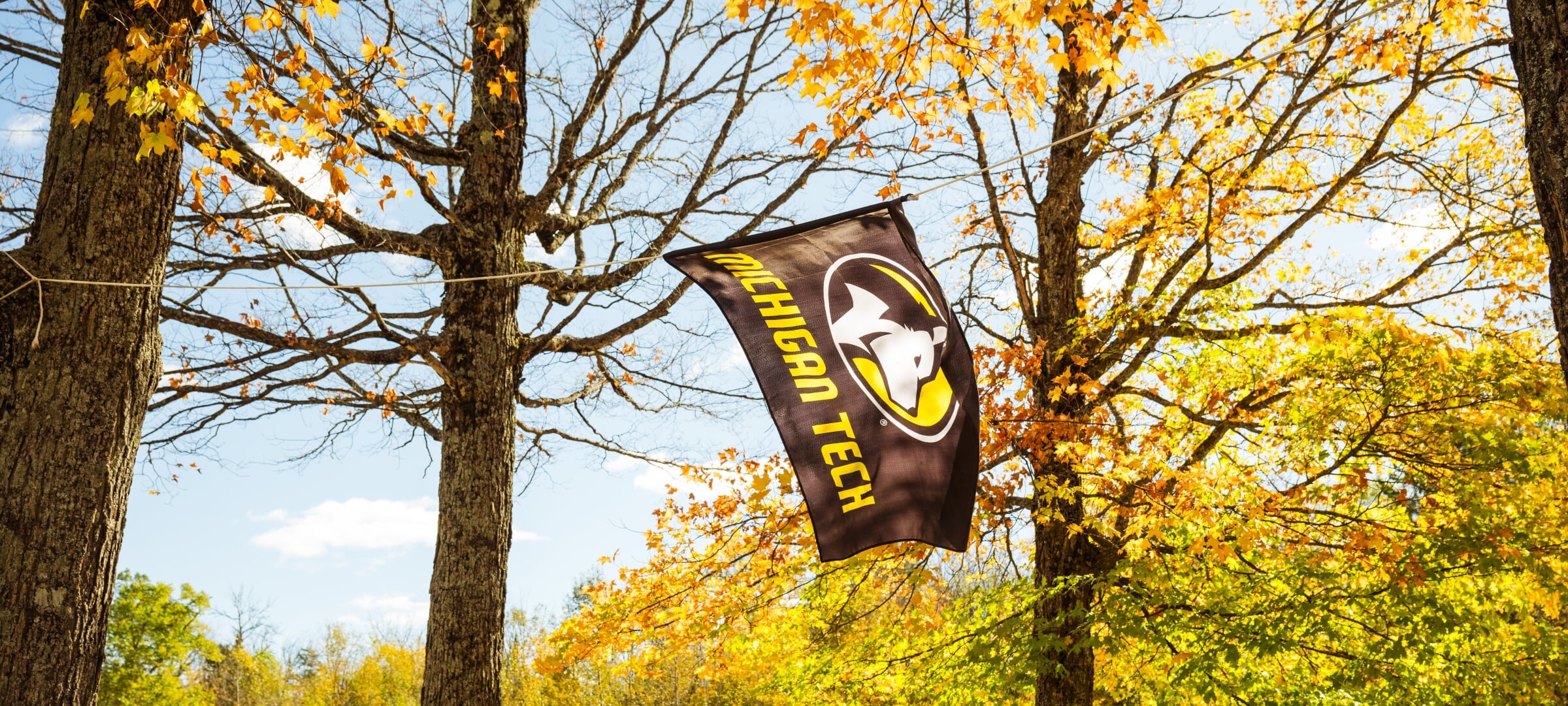 A Michigan Tech flag hun between trees with autumn leaves.