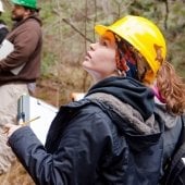 A female student in a hard hat with clipboard looking up in the woods