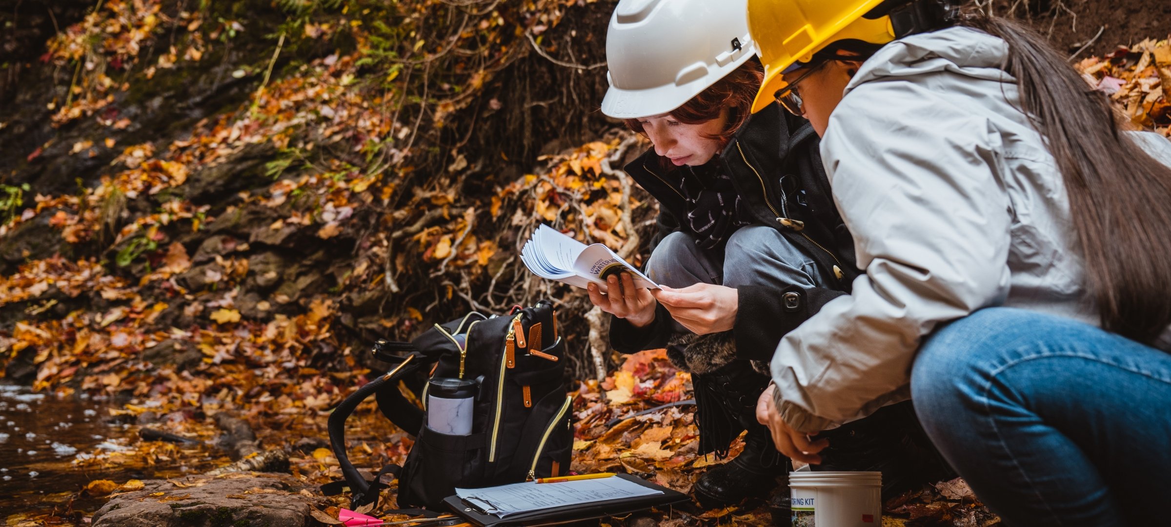 Researchers with hard hats crouching down by a stream looking at a booklet in the woods during fall.