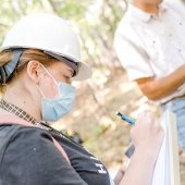 Student with hard hat and mask writing on a clipboard in the woods.