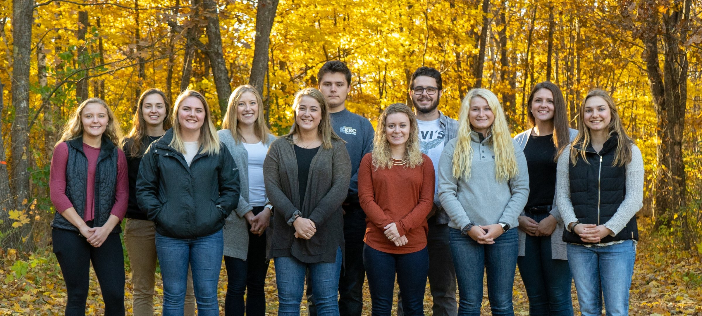 Eleven young people stand in two rows in a fall forest outside smiling and looking at the camera. They are scholarship recipients.