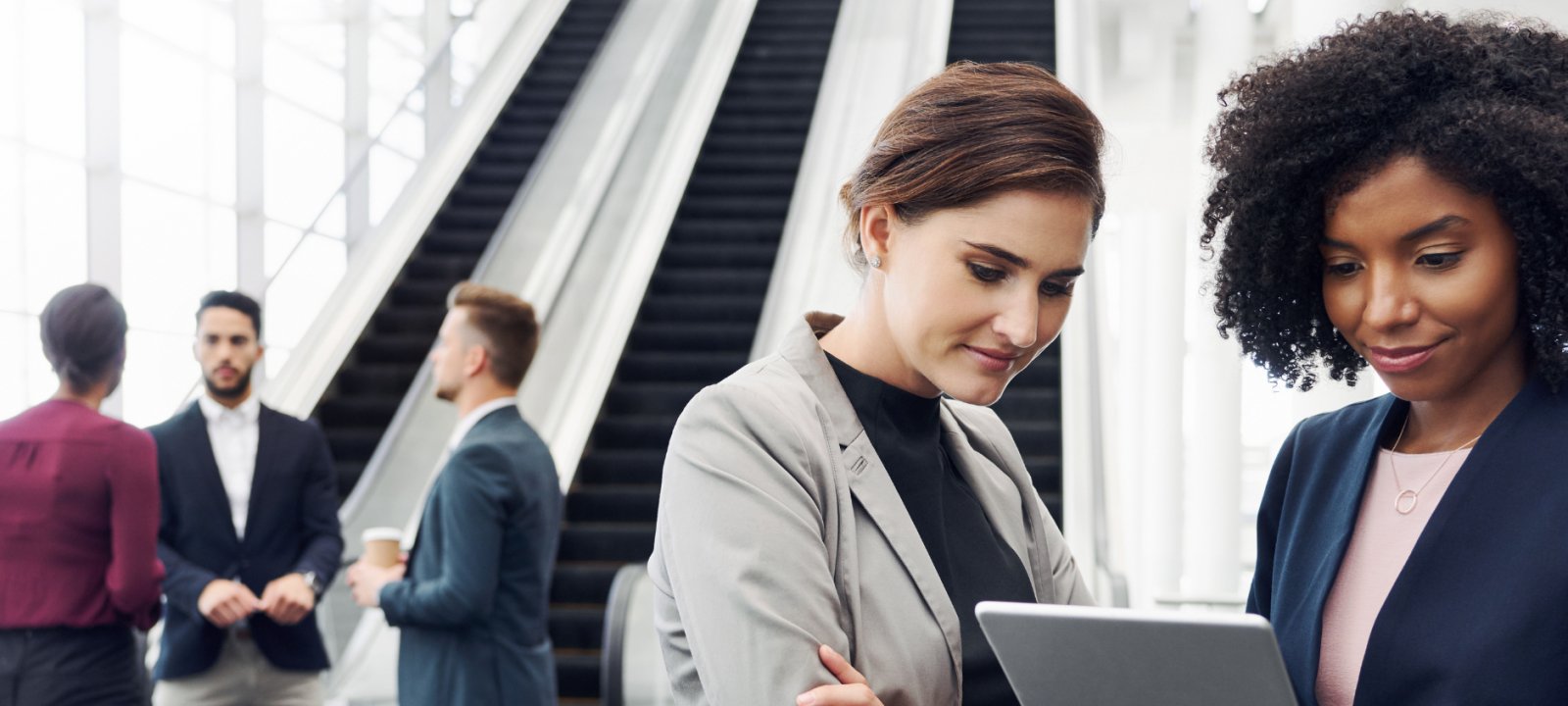 Two standing female business professionals look at a computer while three other employees chat in the background.