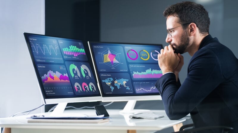 Young man sits in front of several computer monitors looking at severa data visualizations of his business performance.